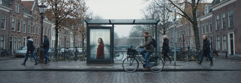 Op een gewone straat hangt in een abri een filmisch affiche van een nieuwe serie; een fietser kijkt even op
